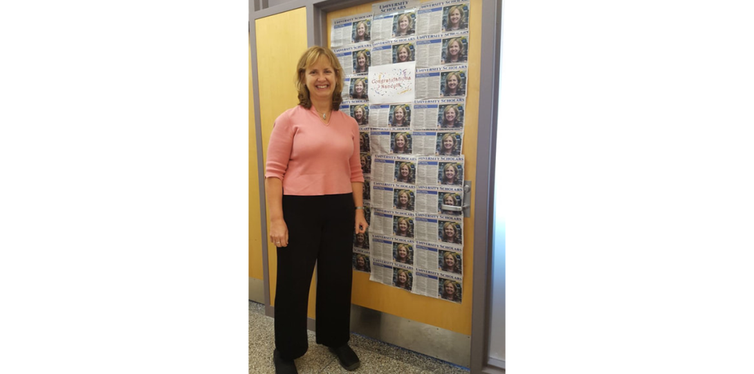Nancy standing next to a board with a multitude of newspaper clippings announcing that she was honored a University Scholar